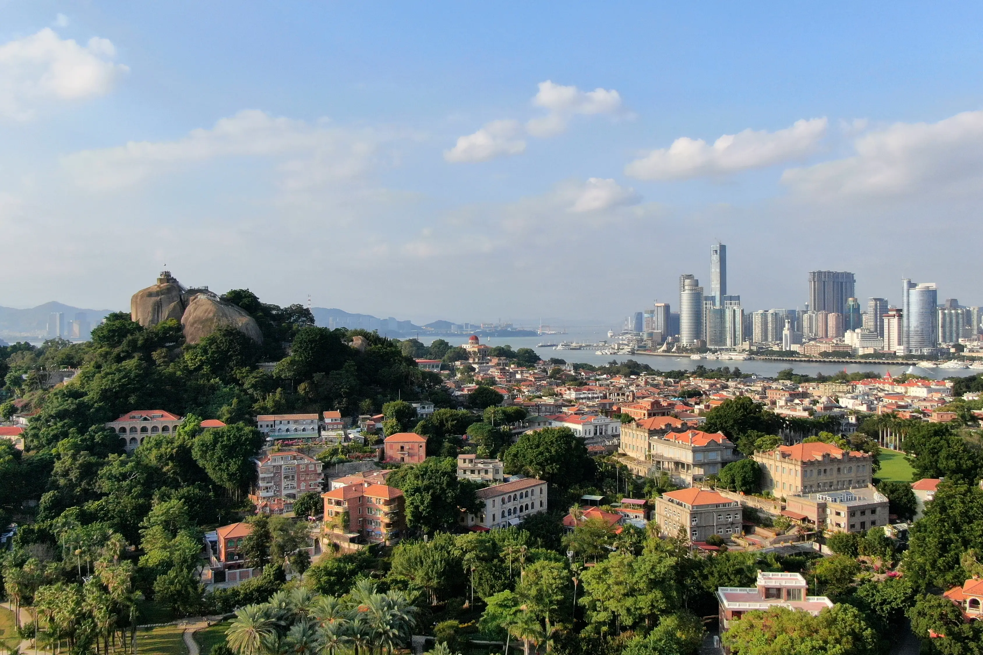 Panoramablick auf Xiamen mit historischen Gebäuden, Felsen und moderner Skyline am Wasser unter blauem Himmel mit weißen Wolken.