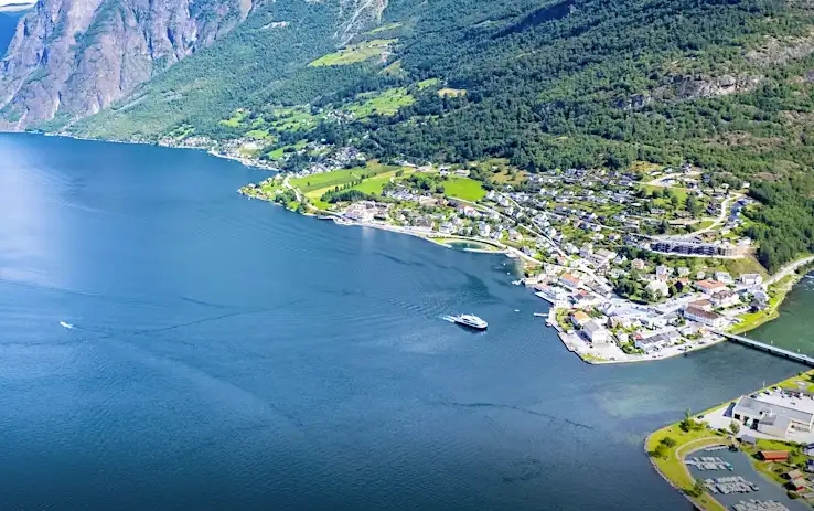 Luftaufnahme eines Küstendorfs, umgeben von Bergen und üppigem Grün, mit einer Fähre, die auf dem tiefblauen Wasser nahe der Küste fährt. Die Landschaft ist geprägt von verstreuten Häusern und Anbauflächen.