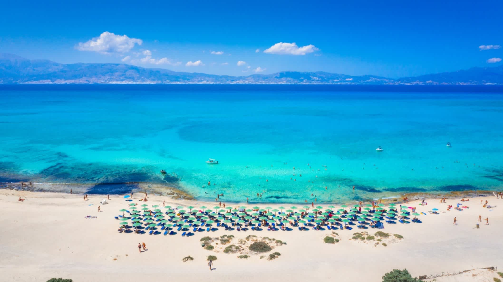 Luftaufnahme des Belegrina Strands auf der Chryssi Insel, Kreta, Griechenland mit azurblauem Meer, Sonnenschirmen und einem Gebirge am Horizont.

