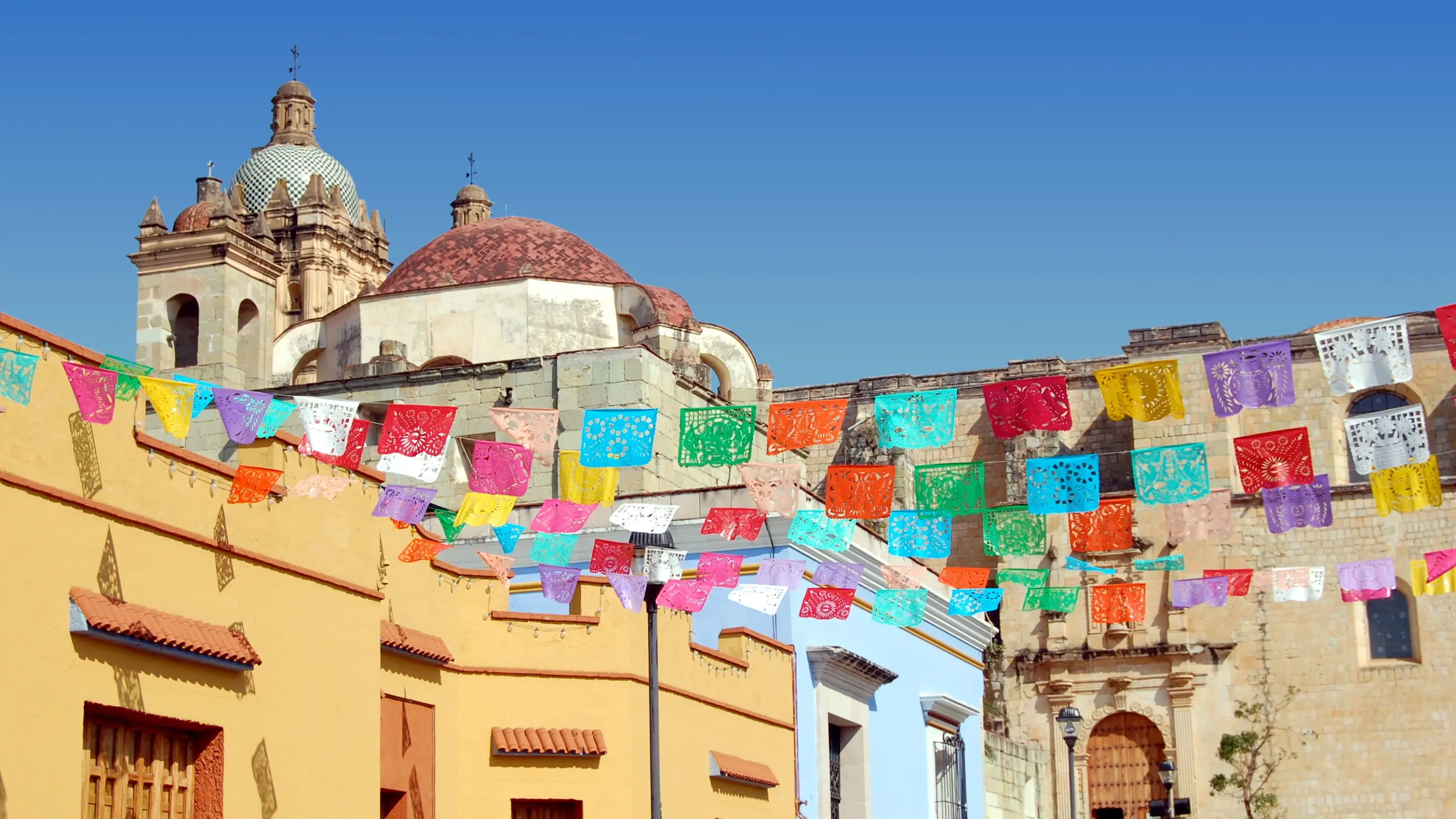 Mexiko, Oaxaca, Gebäude Historische mexikanische Kirche mit Kuppeln unter blauem Himmel, bunte Papierwimpel schmücken die Straße vor gelben Gebäuden.