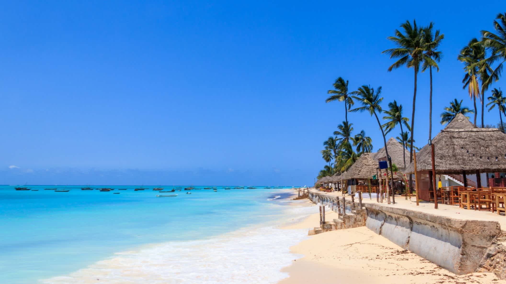 Sandstrand Nunwi Beach in Sansibar mit Pavillons, imposanten Palmen und türkisblauem Meer.
