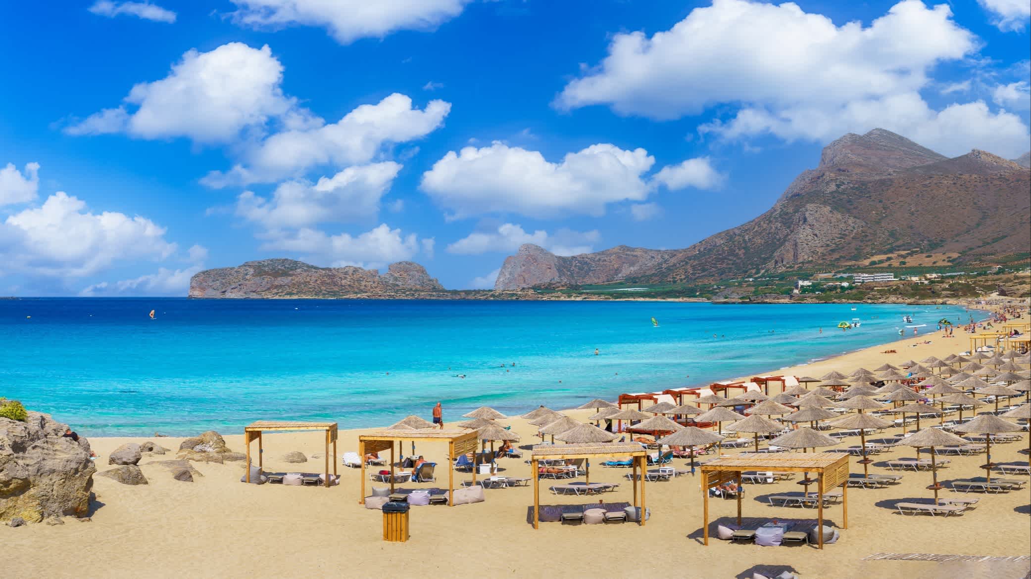 Landschaft mit Falasarna Strand, Kreta, Griechenland mit hellblauem Wasser, vielen Sonnenschirmen und einem Gebirge im Hintergrund.
