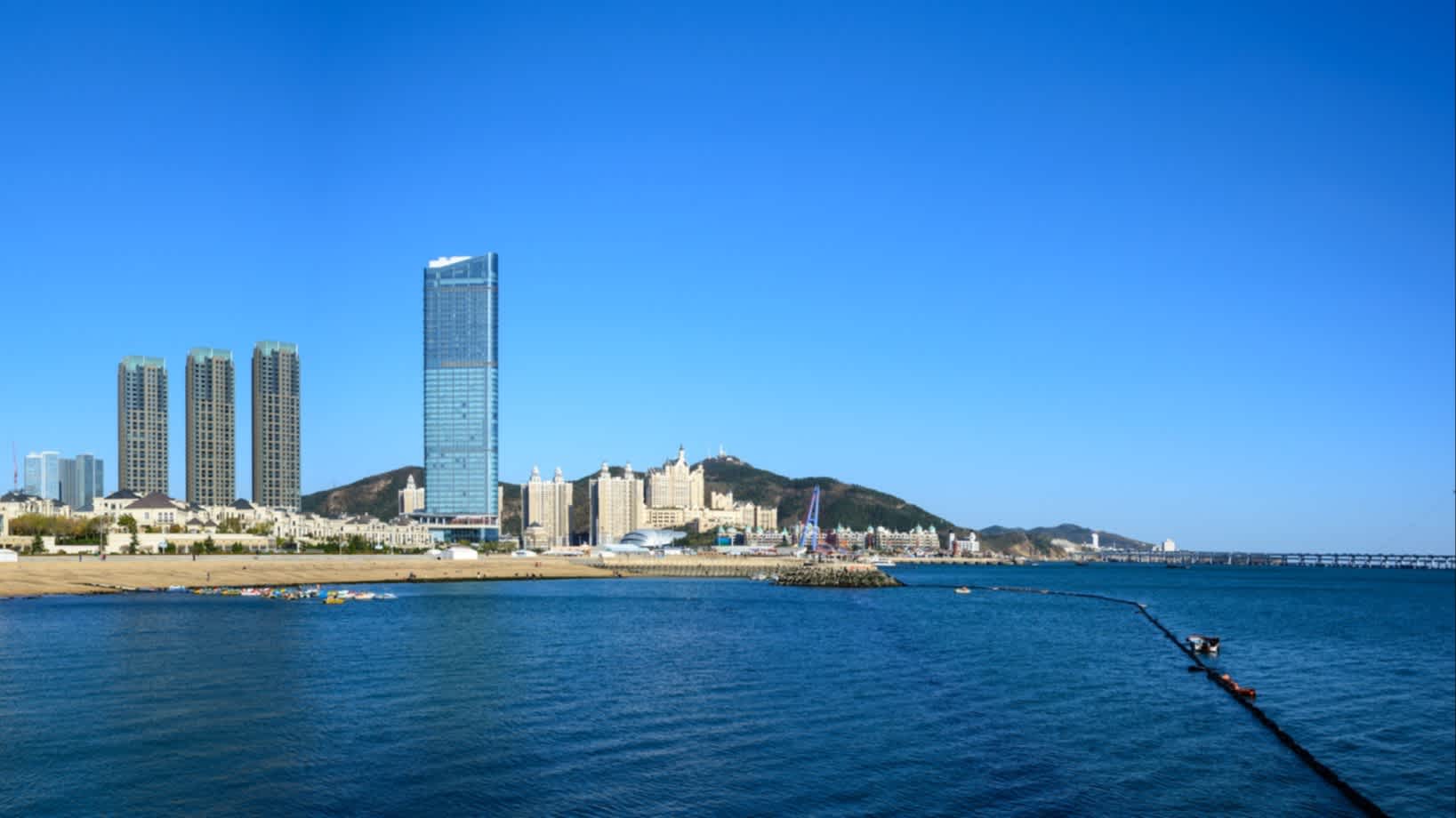 Aussicht auf den Strand und das Meer in Dalian, China bei strahlend Blauem Himmel und mit Hochhäusern im Bild.