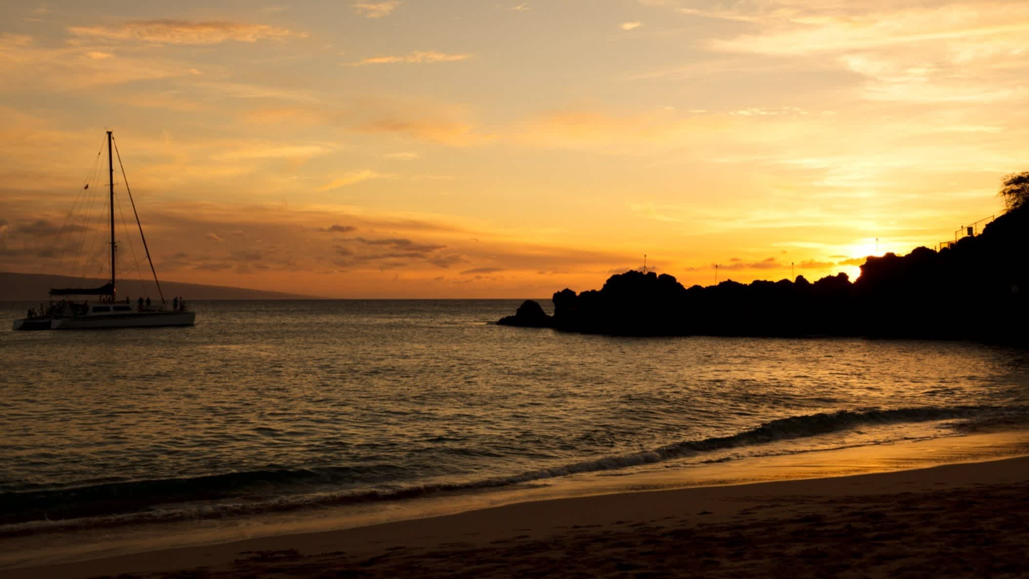 Der Strand Kaanapali-Strand, Maui, Hawaii , USA bei Sonnendämmerung und mit Blick auf das Meer sowie Felsen am Rand des Strandes und einem Seegelboot im Wasser.