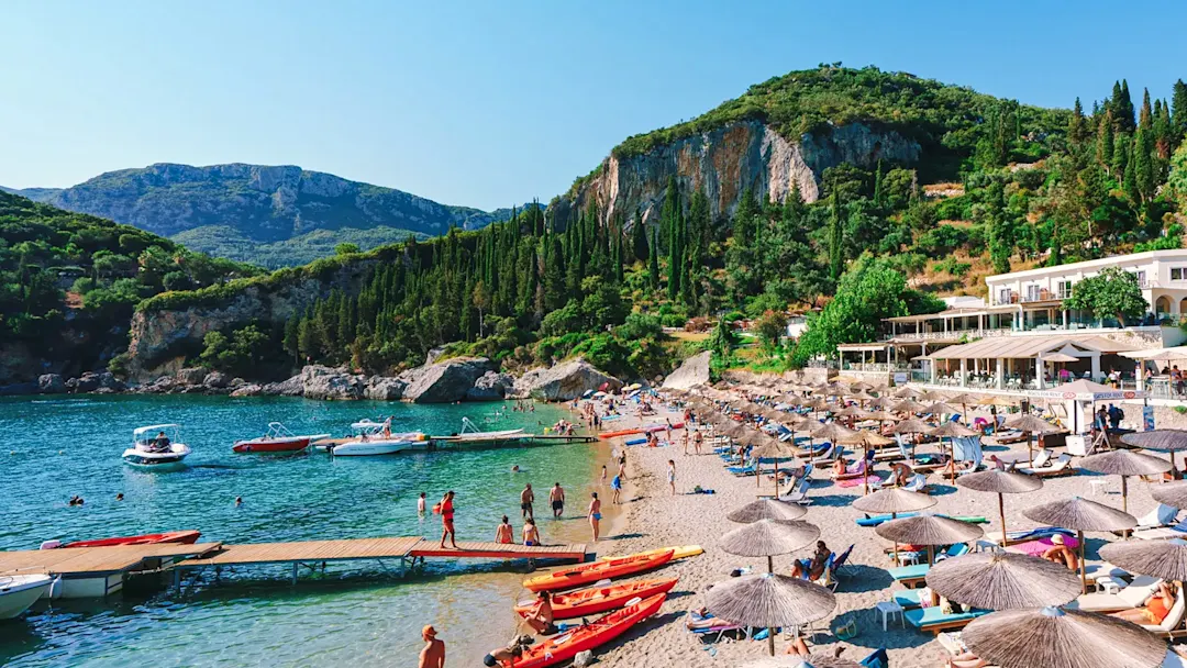 Strand mit Sonnenschirmen, Booten und grünen Hügeln. Paleokastritsa, Korfu, Griechenland.