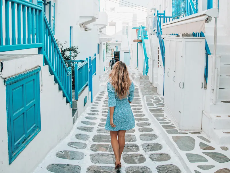 Griechenland, Mykonos, Kykladen, Frau Frau in einem blauen Kleid spaziert durch eine enge, weiße Gasse mit blauen Details. Mykonos, Kykladen, Griechenland.