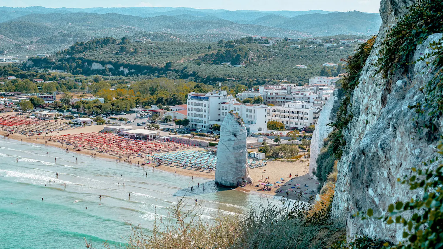 Küstenlandschaft mit türkisblauem Meer, Sandstrand mit bunten Sonnenschirmen, weißer Felsnadel und Hotelanlagen vor grünen Hügeln.