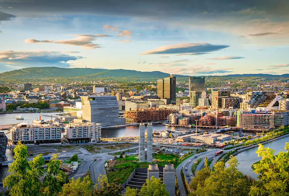 Blick auf eine moderne Stadt mit modernen Gebäuden, Wasserstraßen und grünen Hügeln im Hintergrund bei teilweise bewölktem Himmel, gesehen aus einer erhöhten Perspektive.