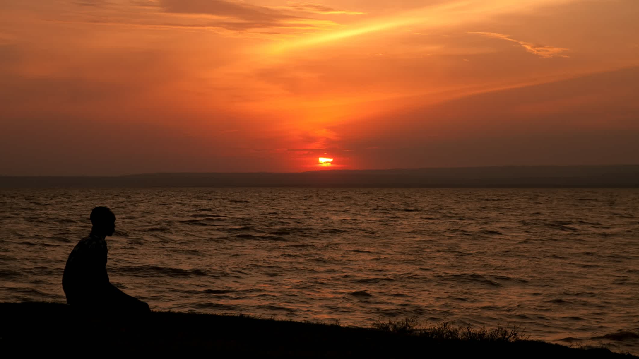 Mann sitzt am Strand und schaut sich den tiefroten Sonnenuntergang am Victoria See an.