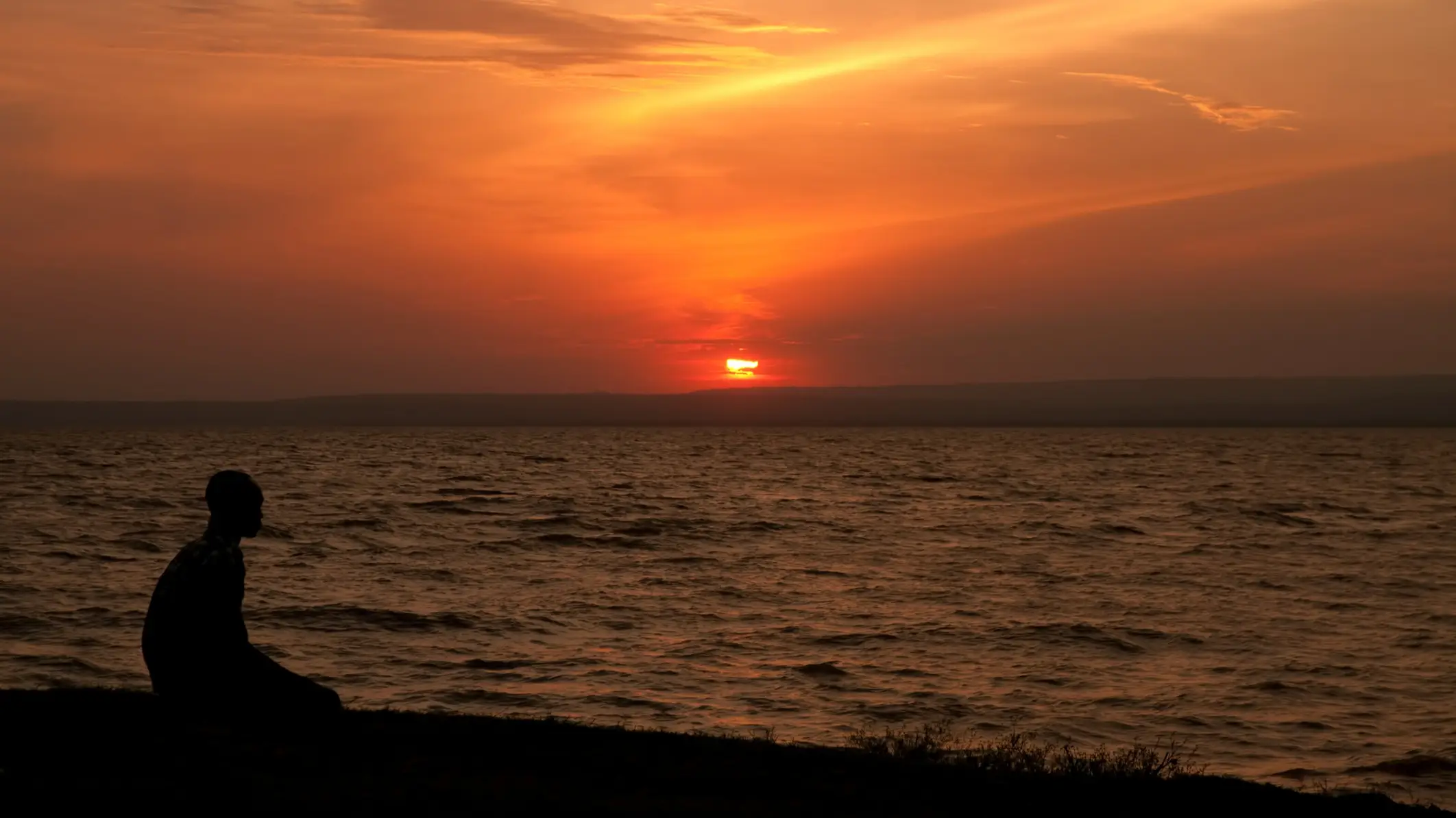 Kenia Lake Victoria Mann sitzt am Strand und schaut sich den tiefroten Sonnenuntergang am Victoria See an.