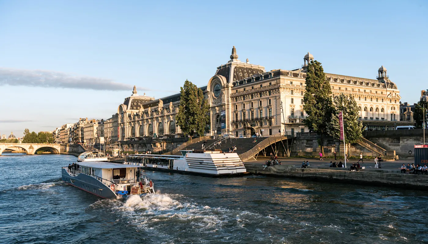 Musée d'Orsay along the Seine River in Paris with tourist boats cruising by under clear blue sky.