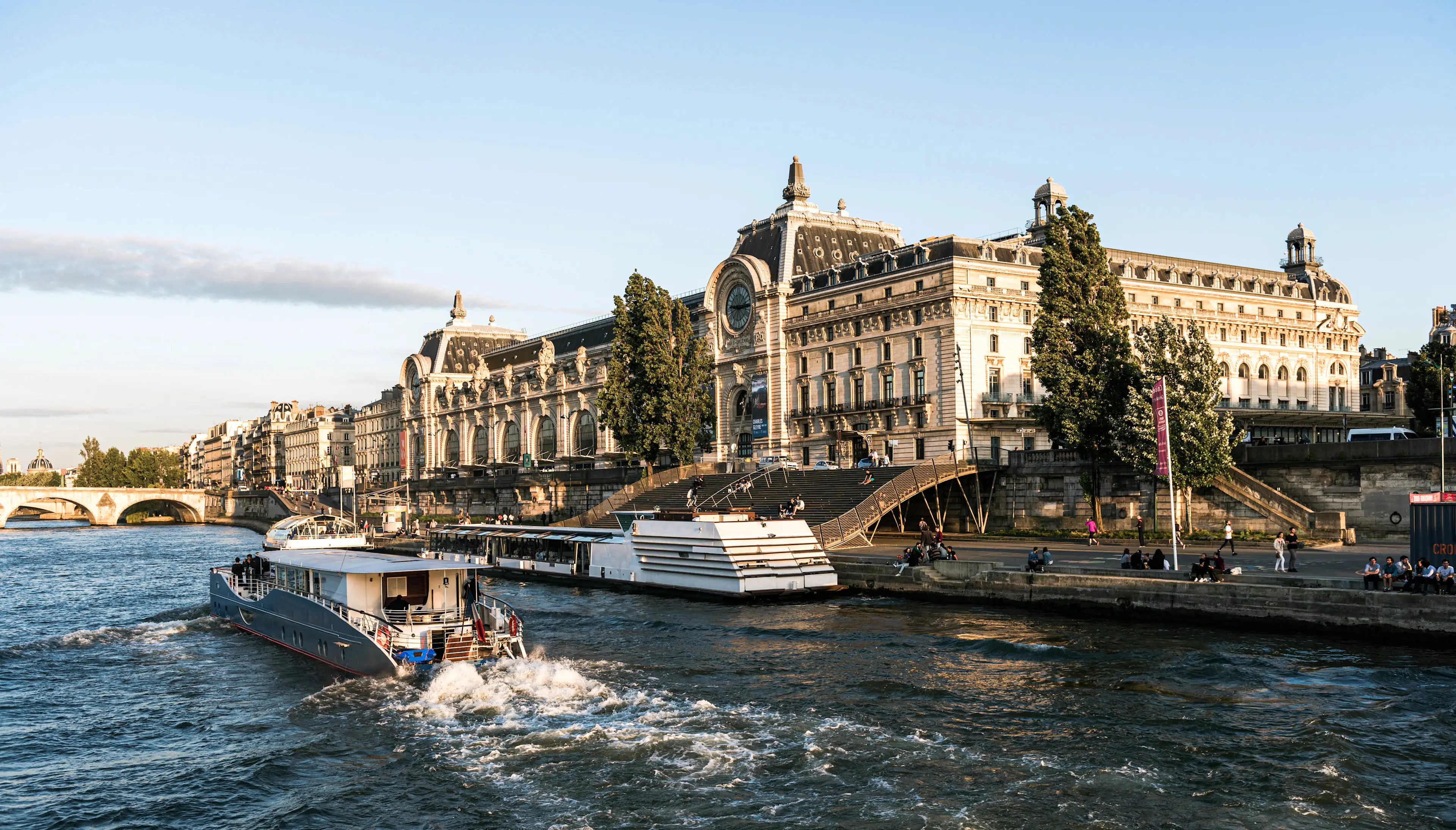 Musée d'Orsay along the Seine River in Paris with tourist boats cruising by under clear blue sky.