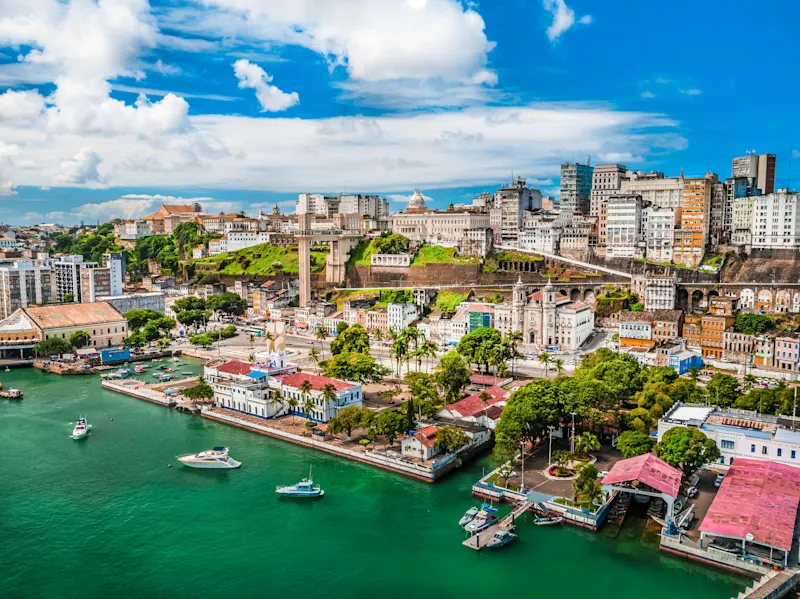 Luftaufnahme von Salvador da Bahia mit türkisblauem Hafen, historischer Altstadt auf dem Hügel und blauem Himmel mit Wolken.