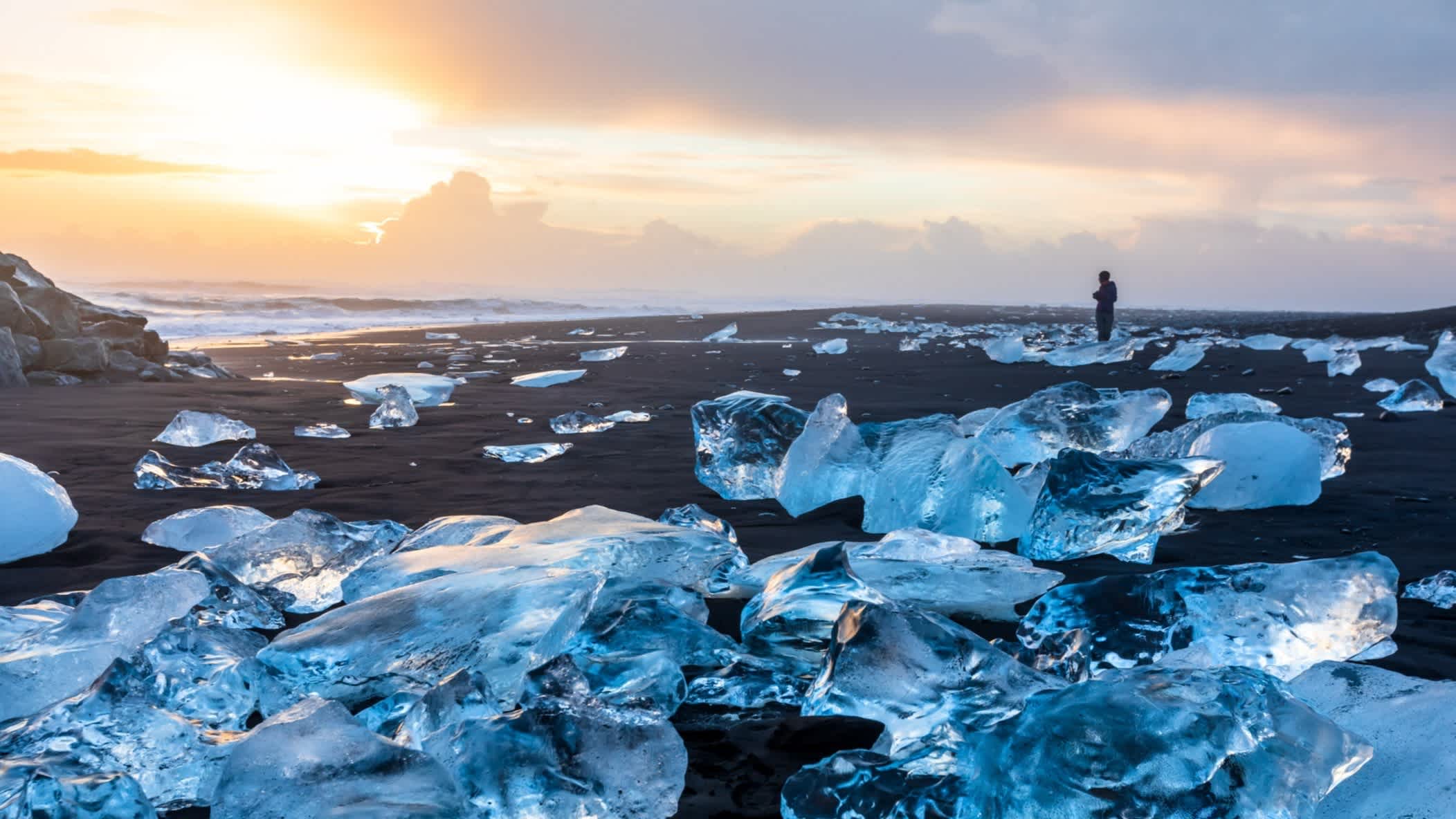 Diamond Beach in Jokulsarlon, Island, bei Sonnenuntergang und mit blauen Eiskristallen, die auf dem schwarzen Sandstrand glitzernd funkeln.
