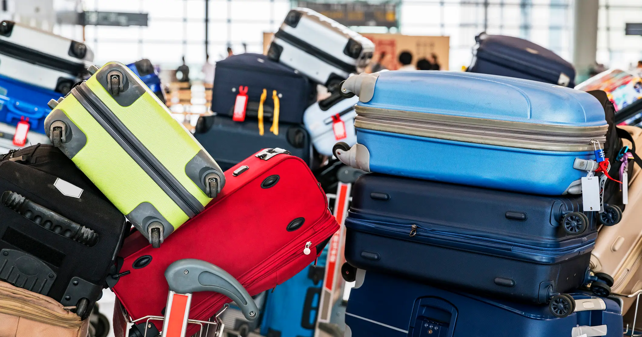 Mehrere bunte Koffer, darunter rote, blaue, grüne und schwarze, stapeln sich wahllos auf Gepäckwagen in einem Flughafenterminal mit verschwommenem Hintergrund.