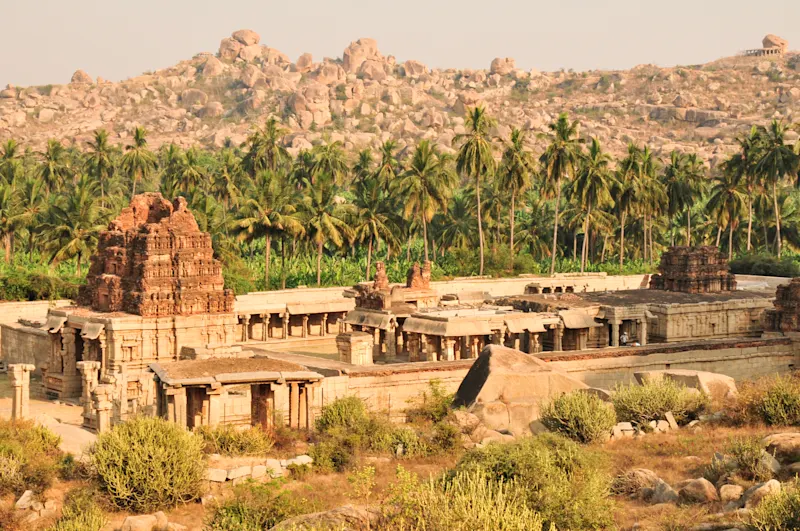 Antiker Tempel in Hampi mit Palmenreihe und felsiger Berglandschaft im Hintergrund bei goldenem Sonnenlicht.