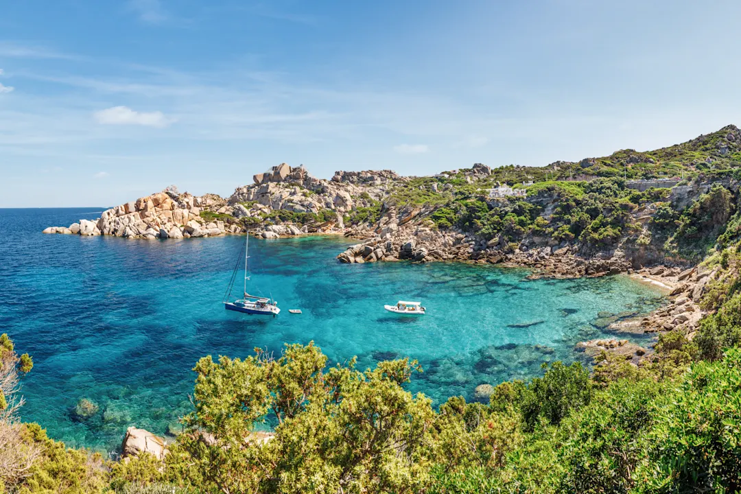 Klare, türkisfarbene Bucht mit Felsen und grünem Ufer bei Sardinien, Italien.