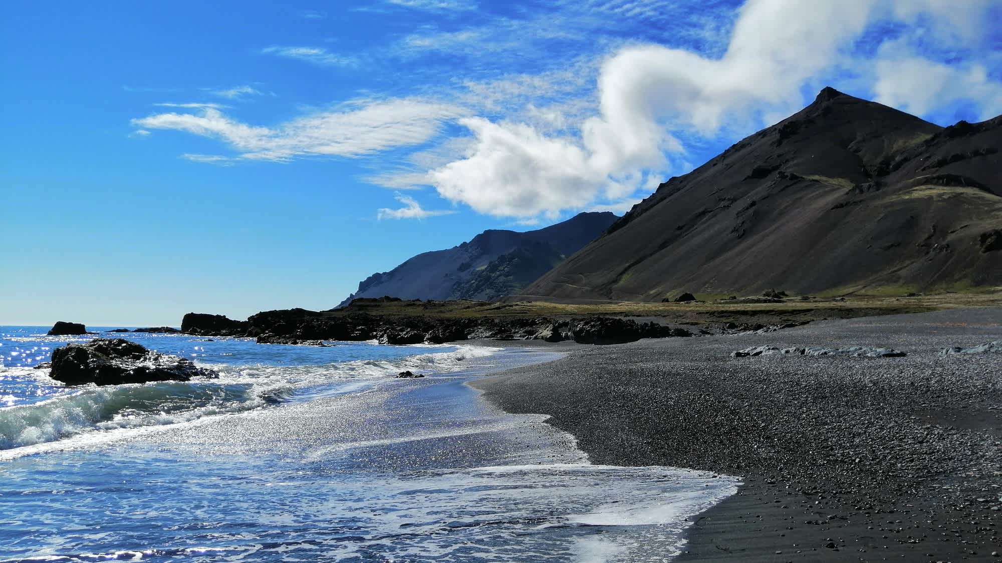Blick auf den schwarzer Sandstrand von Fauskasandur in Island bei blauem Himmel und mit traumhaften Bergen im Hintergrund.