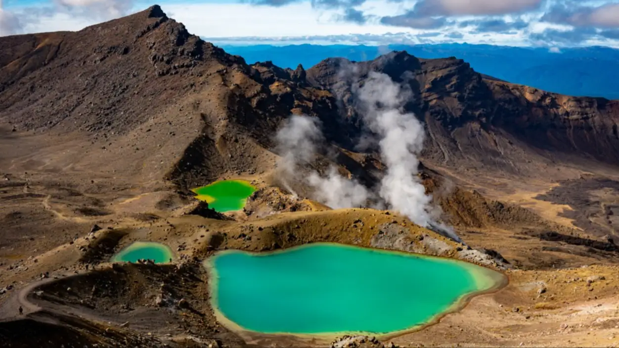 Vulkanische Landschaft mit türkisfarbenen Kraterseen und aufsteigendem Dampf unter blauem Himmel mit Wolken.