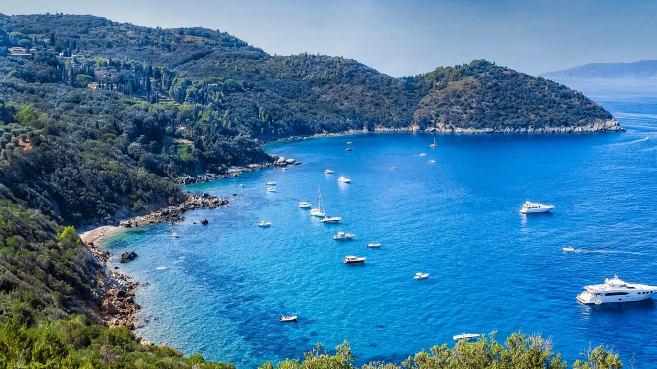 Luftaufnahme des Golfs von Baratti, Toskana, Italien mit Blick auf grüne Hügel sowie Booten im Meer.

