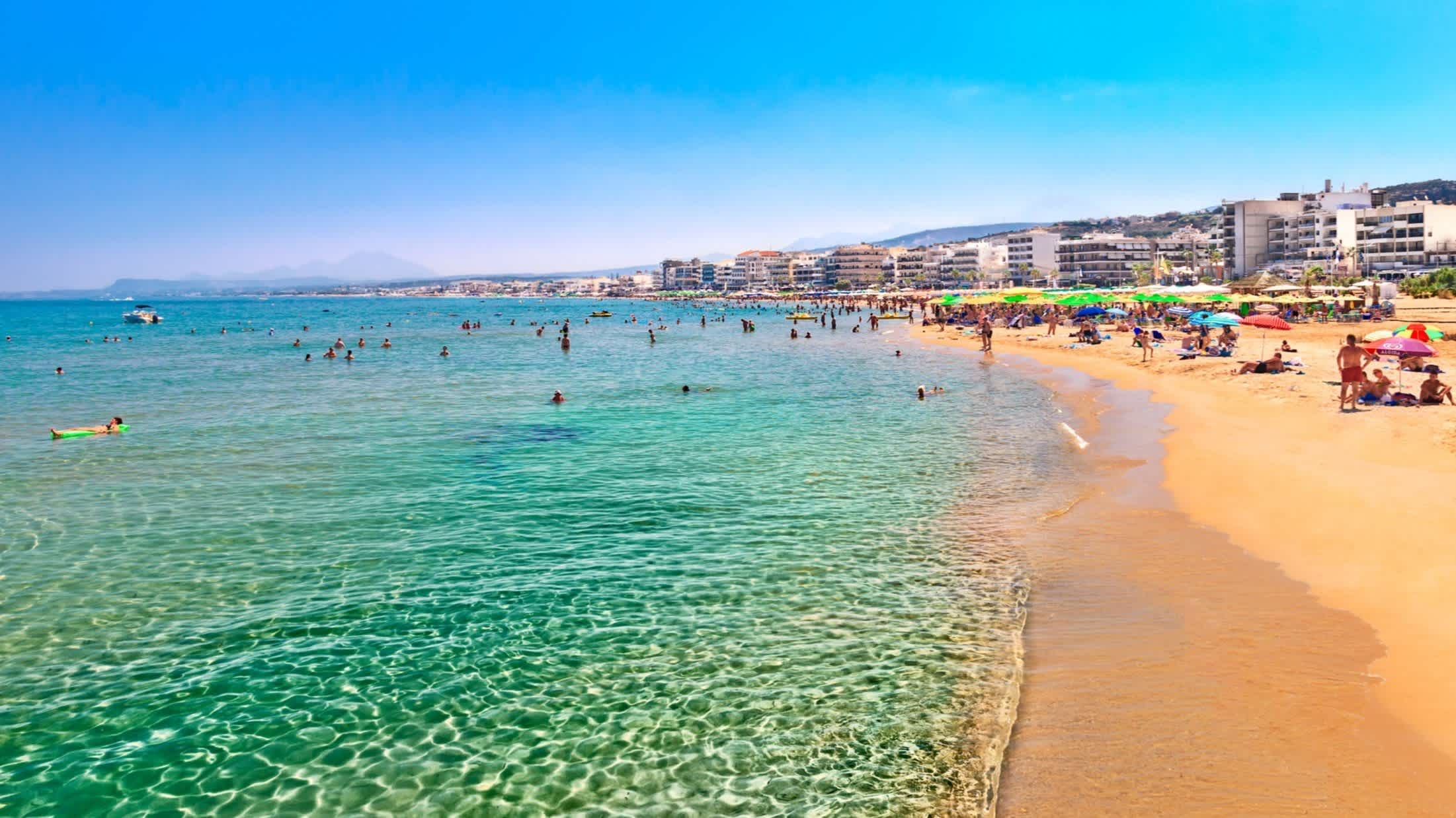 Blick zum Stadtstrand von Rethymno, Kreta. Griechenland bei klarem grünem Wasser und mit vielen Menschen am Strand.