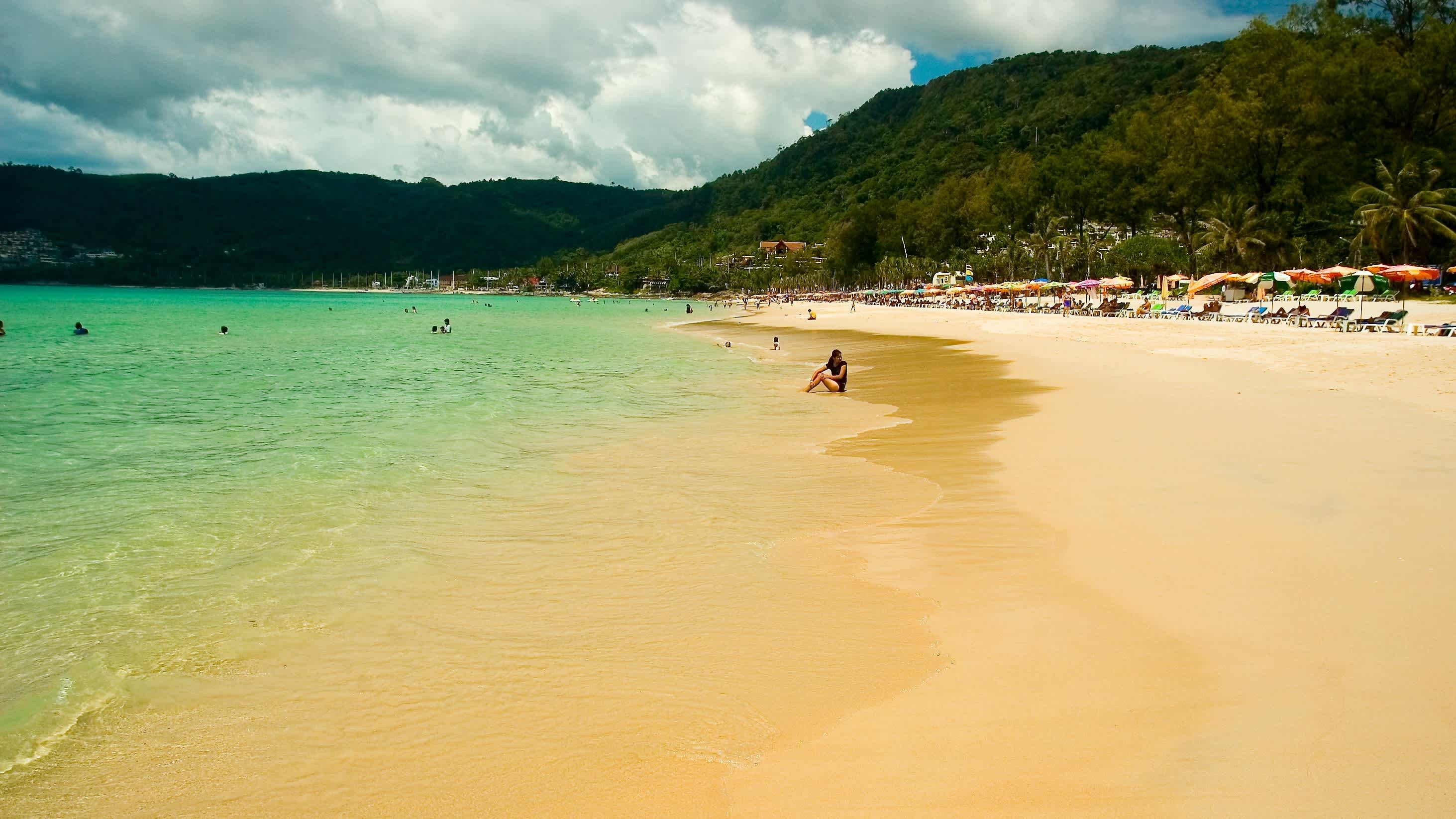 Der Patong Beach auf Phuket, Thailand, mit goldgelbem Sand, seichtem, türkisblauem Meer und bunten Sonnernschirmen im Hintergrund.