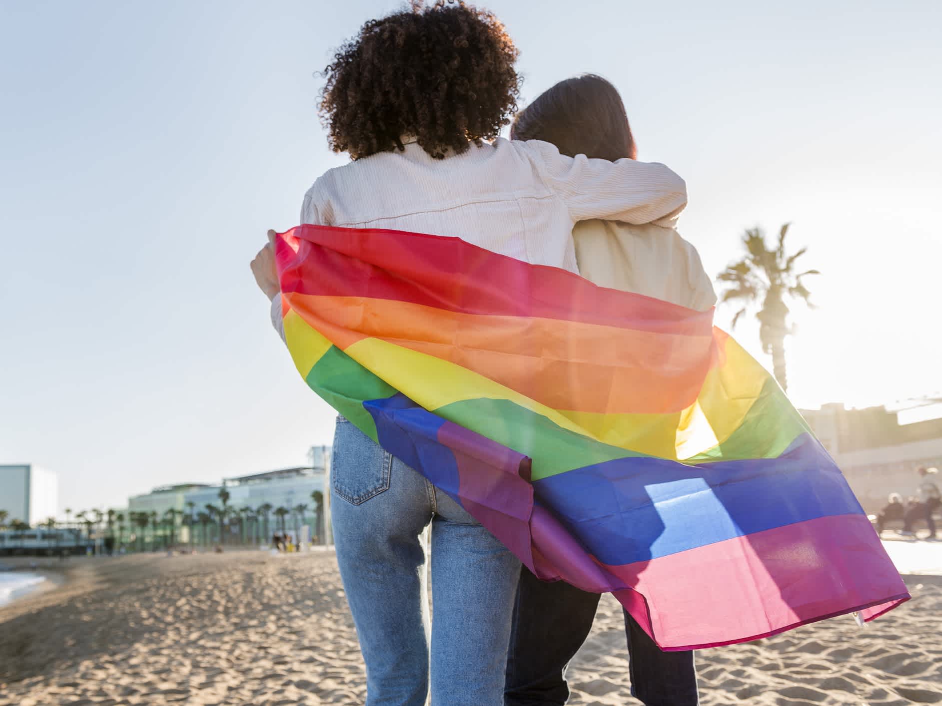 Ein Paar mit der Regenbogen-Flagge auf dem Barceloneta-Strand, Katalonien, Spanien. 