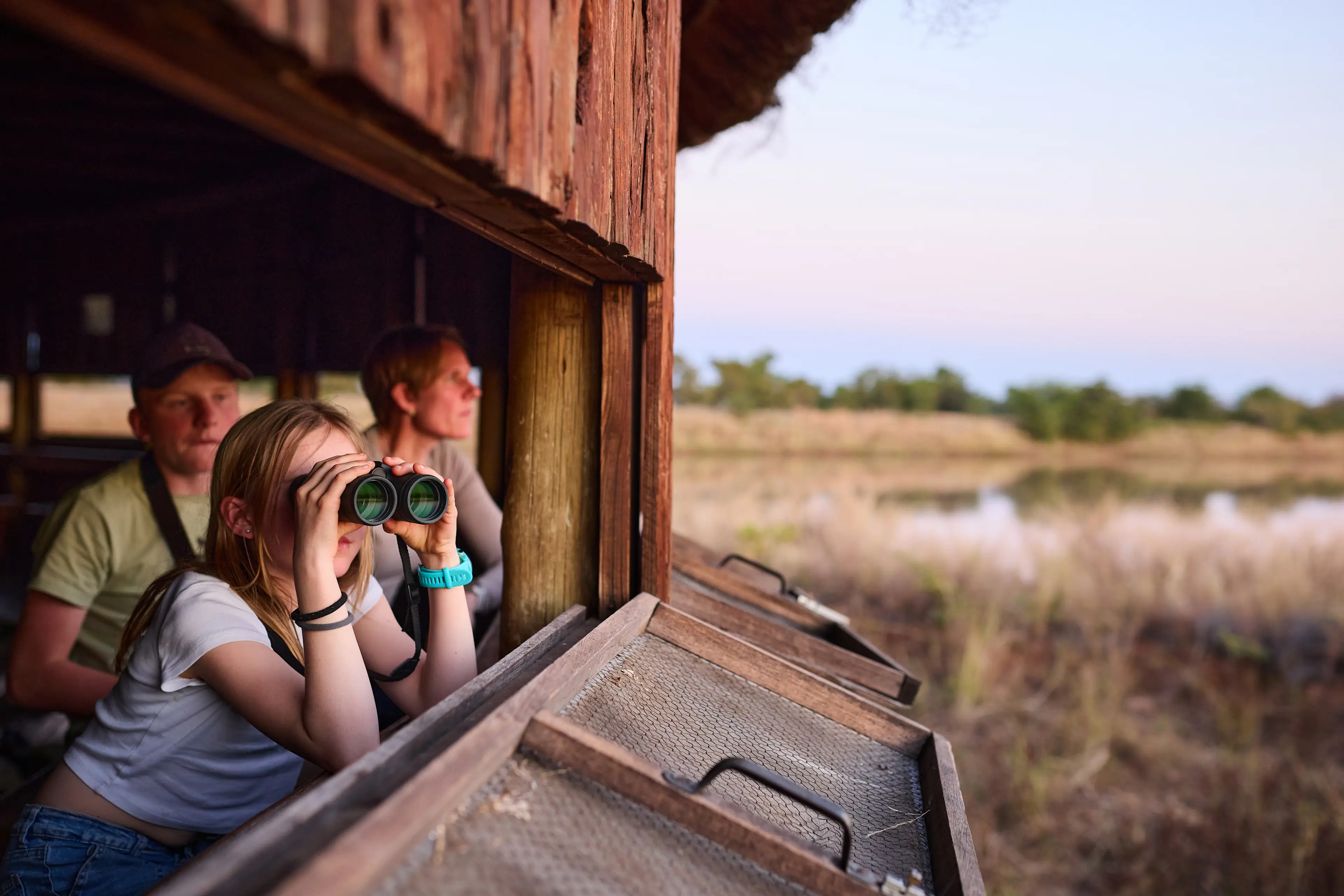 Botswana, Familienreise Personen in einem Holzbeobachtungsstand blicken mit Fernglas auf eine Naturlandschaft mit Wasserfläche in der Dämmerung.
