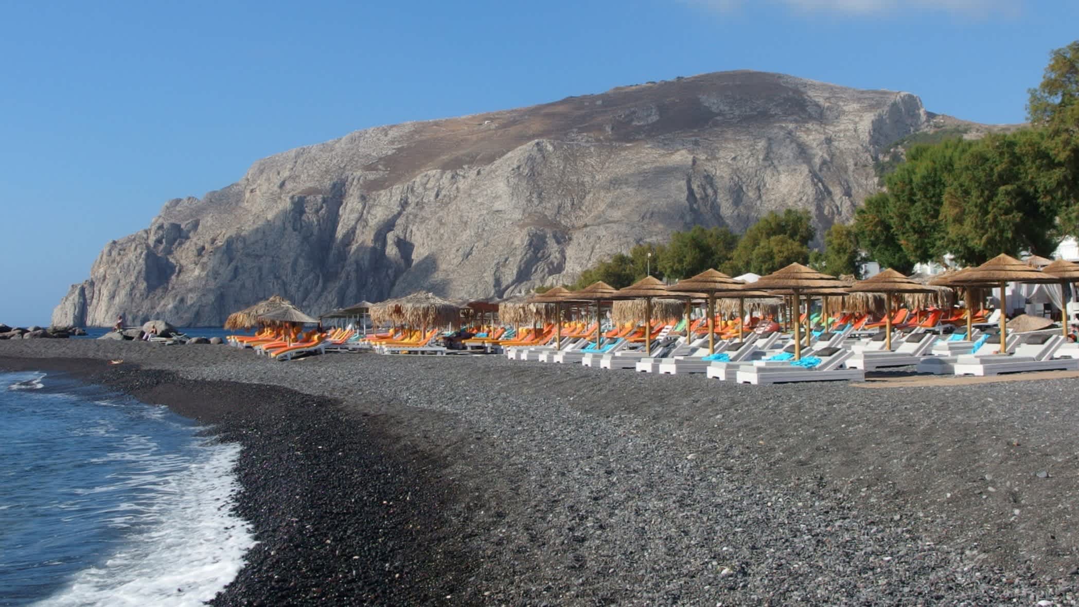 Strand in Kamari an einem Sommertag, Santorini, Griechenland und mit einem Gebirge im Hintergrund sowie Sonnenschirmen am schwarzen Strand.