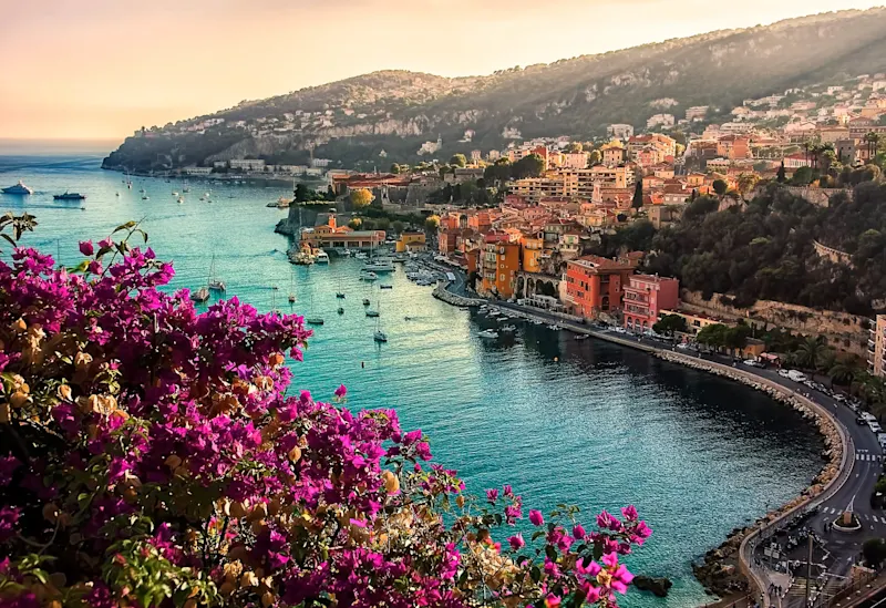 Sunset view of French Riviera coastline with vibrant pink bougainvillea flowers, turquoise water, and colorful hillside buildings.
