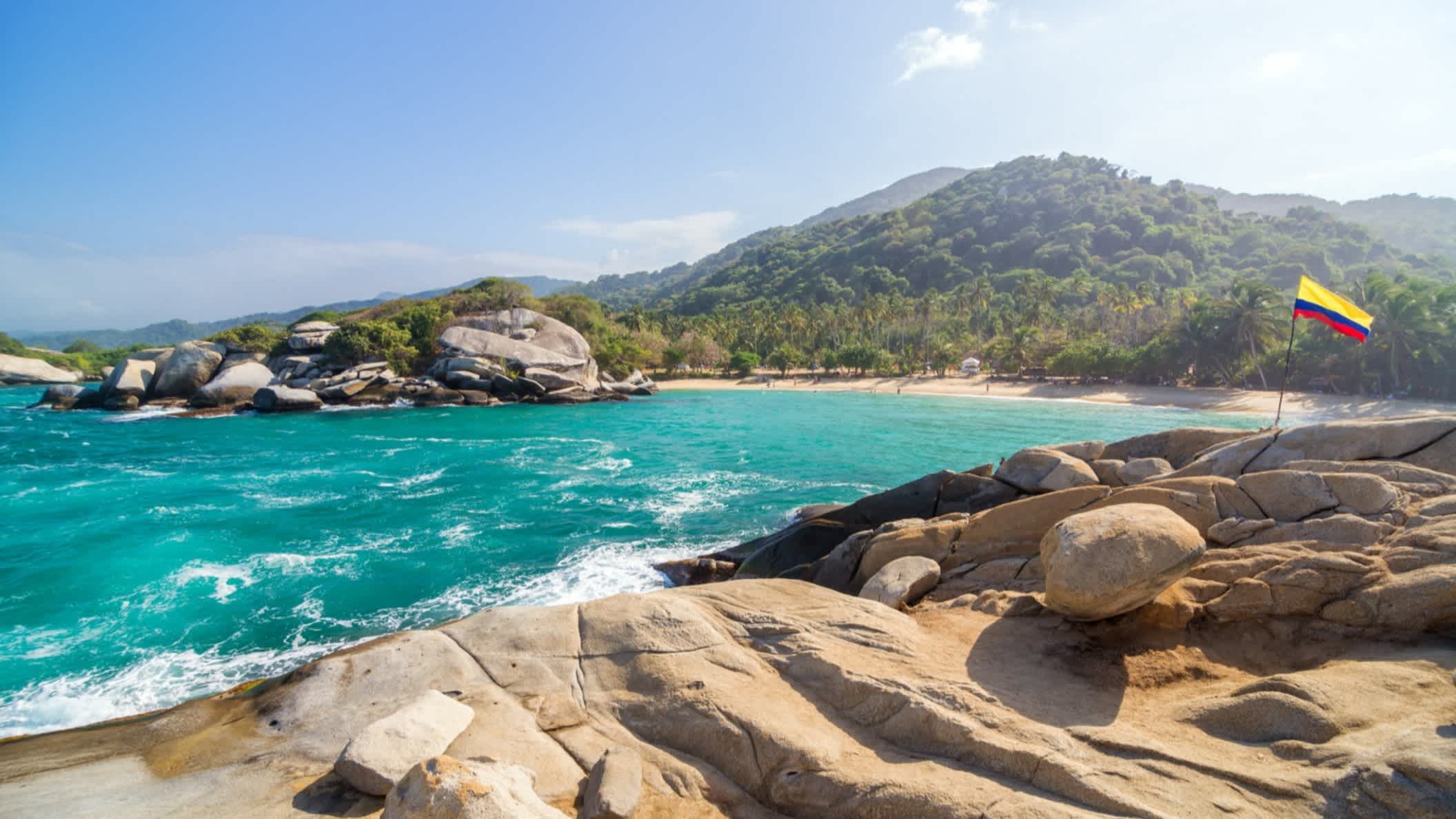 Tropischer Strand im Tayrona-Nationalpark in Kolumbien mit einer kolumbianischen Flagge im Hintergrund sowie Felsen, dem Meer und Bergen.