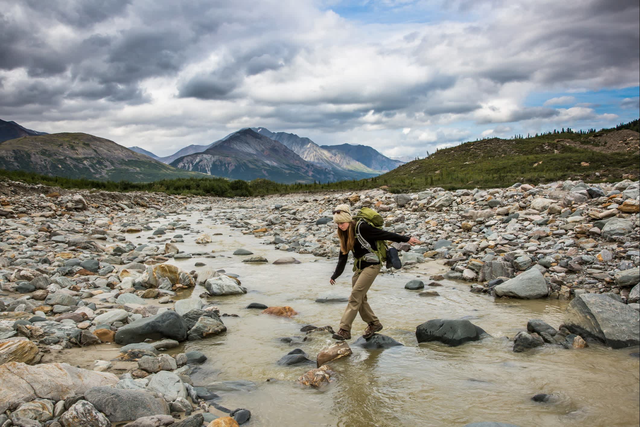 Rucksacktouristin balanciert auf Felsen und überquert einen Fluss im Wildnisgebiet, Alaska.