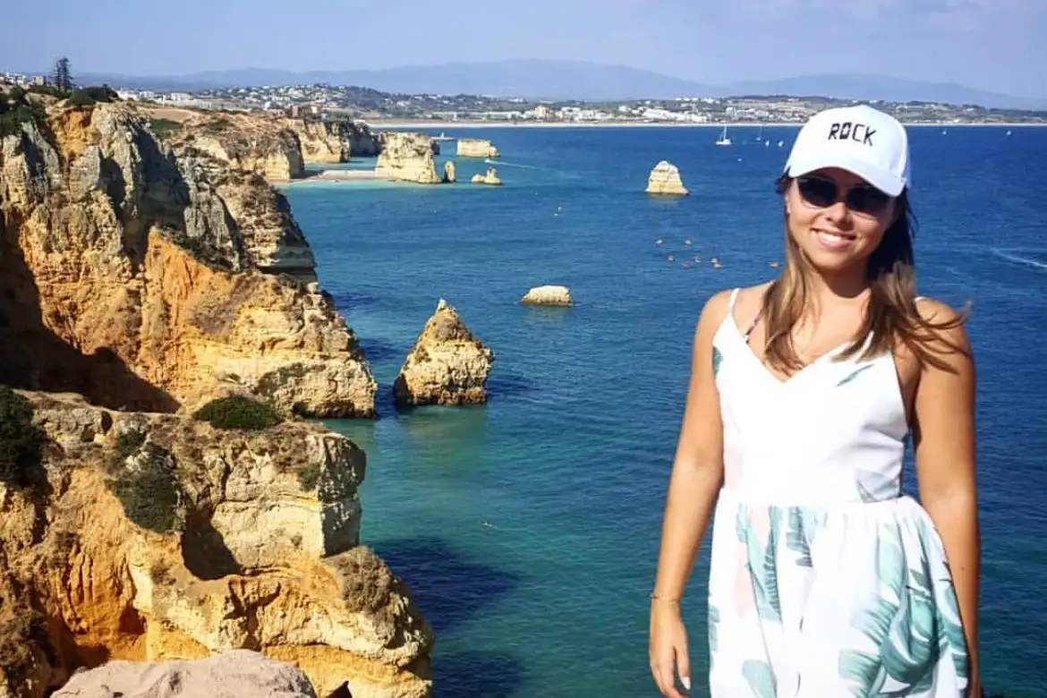 Raphaela, en robe blanche et casquette devant les falaises dorées et l'eau turquoise de l'Algarve au Portugal.