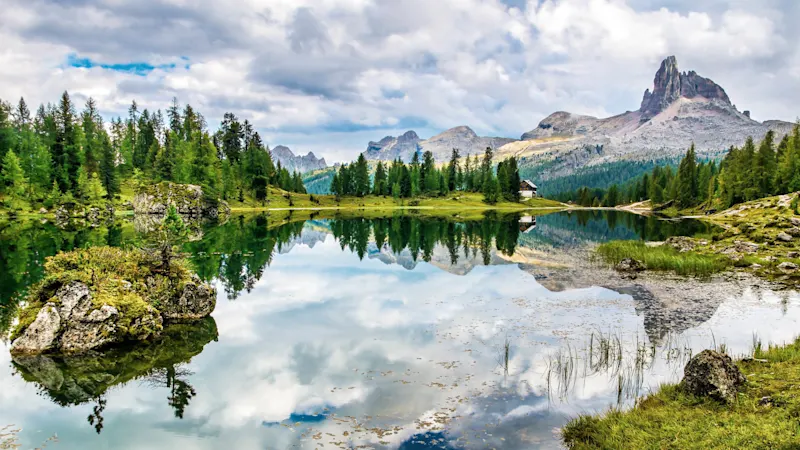 Serene alpine lake perfectly reflecting jagged mountain peaks, evergreen forests, and cloudy sky with a small cabin visible.