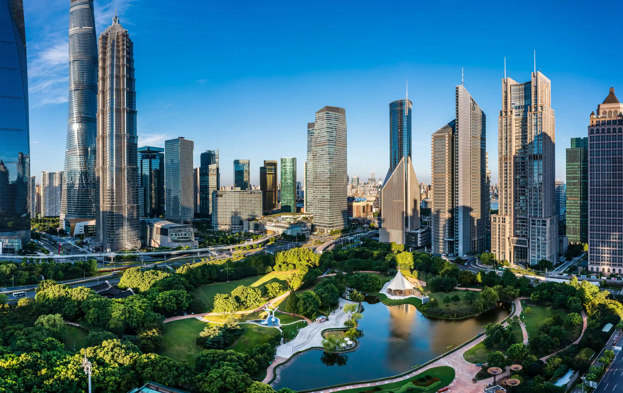 Moderne Skyline von Shanghai mit Wolkenkratzern und grünem Stadtpark mit See unter blauem Himmel.