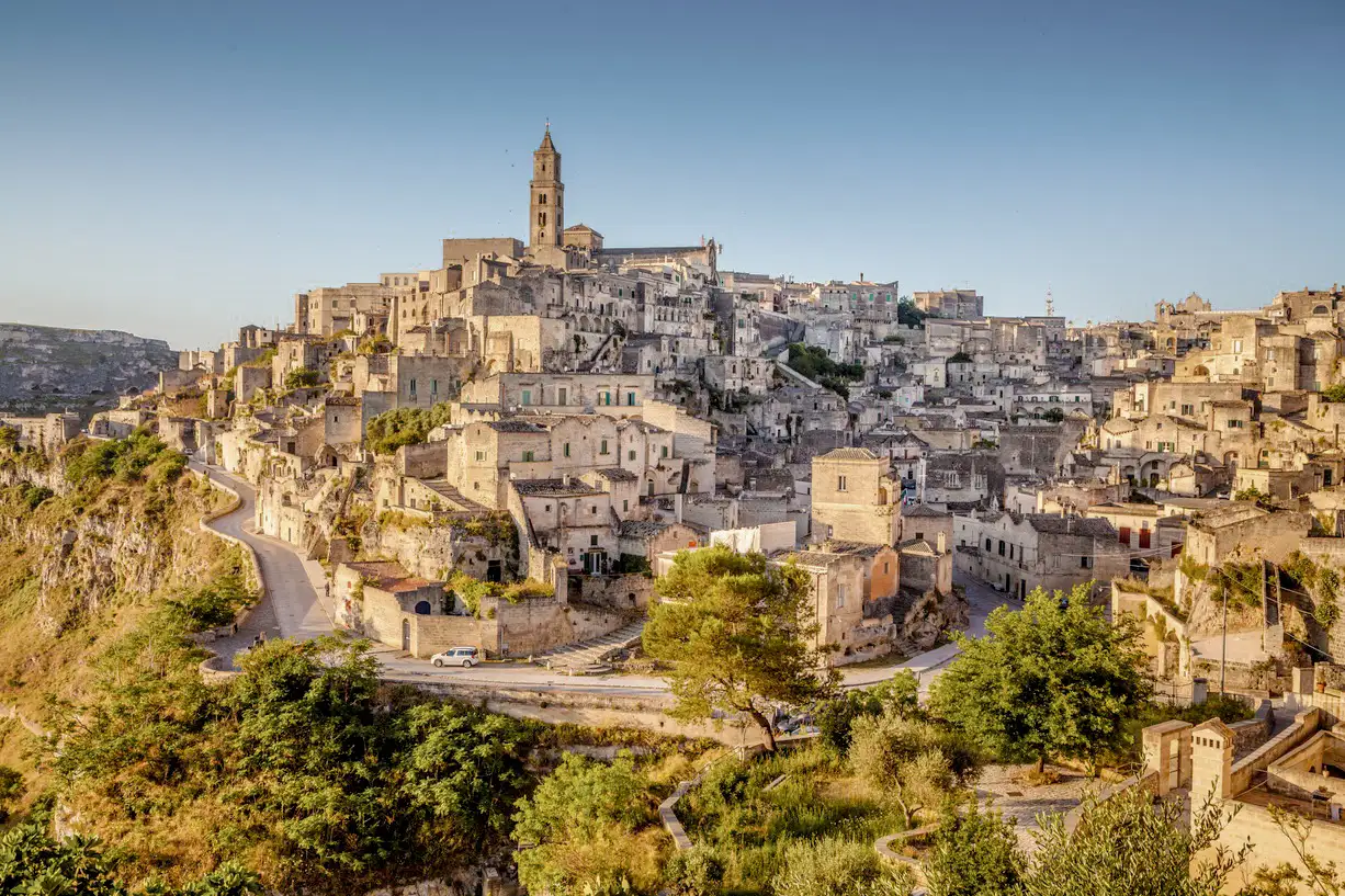 Historische Stadt auf einem Hügel mit steinernen Gebäuden und einem zentralen Kirchturm, umgeben von Grün und felsigen Klippen unter einem klaren blauen Himmel, der in warmes Sonnenlicht getaucht ist.