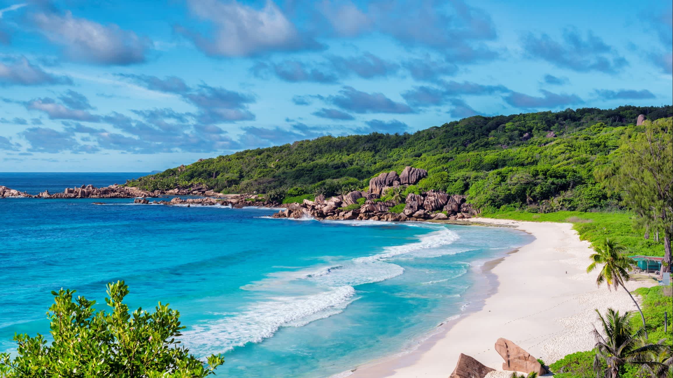 Blick von einem Aussichtspunkt aus auf den Strand Grand Anse auf den Seychellen, Insel La Digue bei herrlichem Sonnenschein und mit azurblauen Wellen und weißem Sand sowie üppiger Vegetation und Bergen.