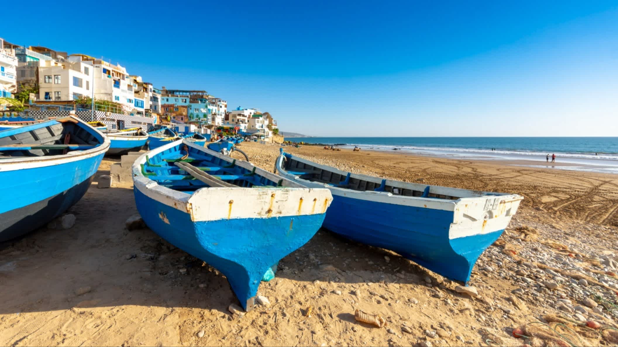 Der Strand von Taghazout, Marokko mit Blick auf blaue Boote am Strand, Menschen sowie Häusern entlang der Promenade. 