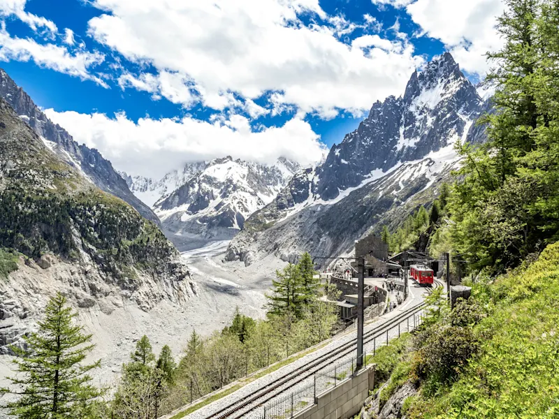 Red mountain train on tracks with alpine station surrounded by snow-capped peaks and green forest under blue sky.