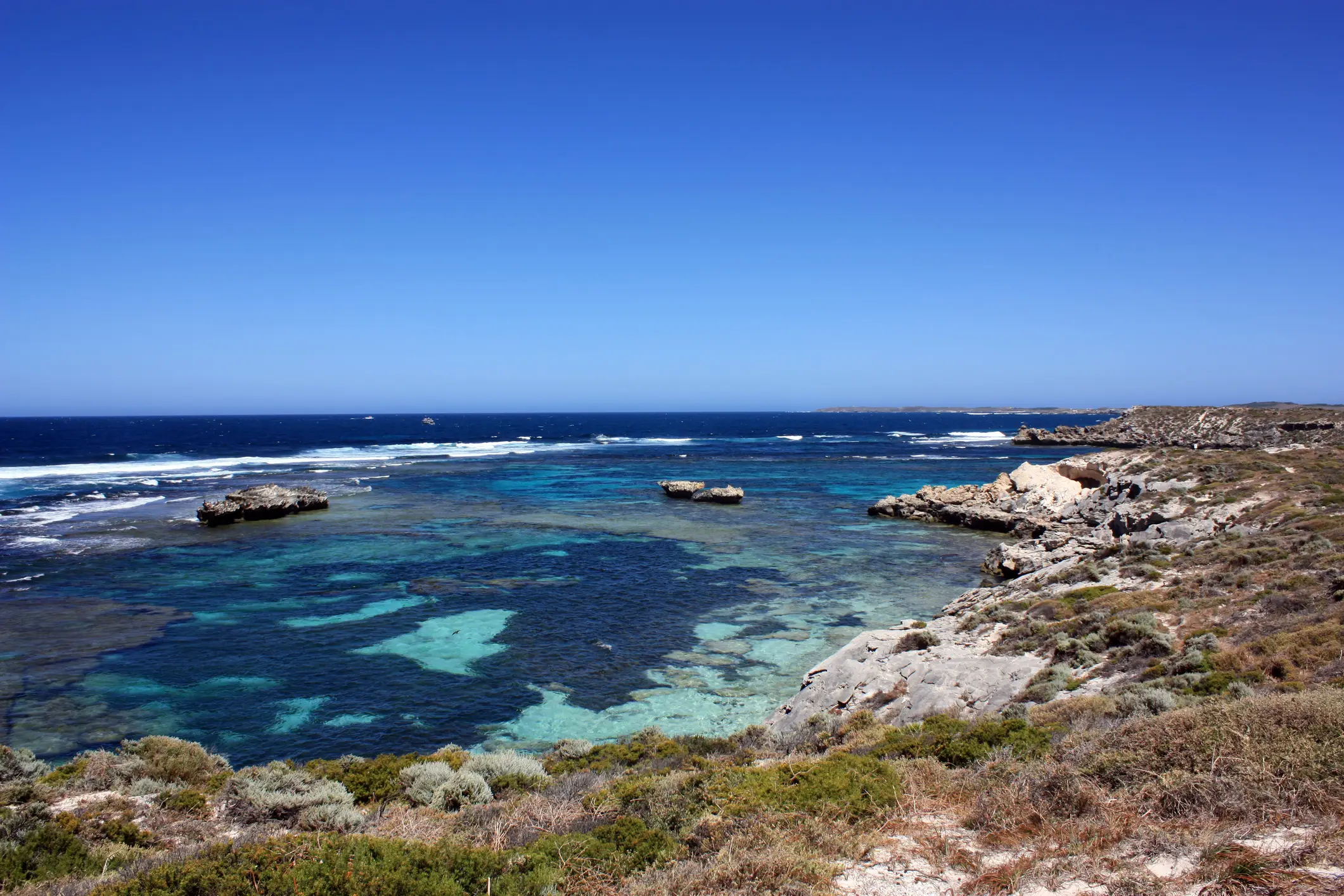 Eine felsige Küste mit kristallklarem Wasser, türkisfarbenen Riffen und sanften Wellen unter einem strahlend blauen Himmel. Die Küstenvegetation bedeckt den Vordergrund und der Horizont erstreckt sich weit in die Ferne.