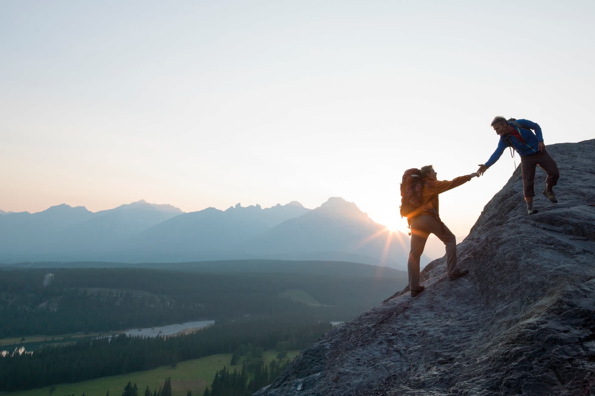 Zwei Bergsteiger bieten helfende Hand auf einem Felsrücken bei Sonnenaufgang über einem Tal