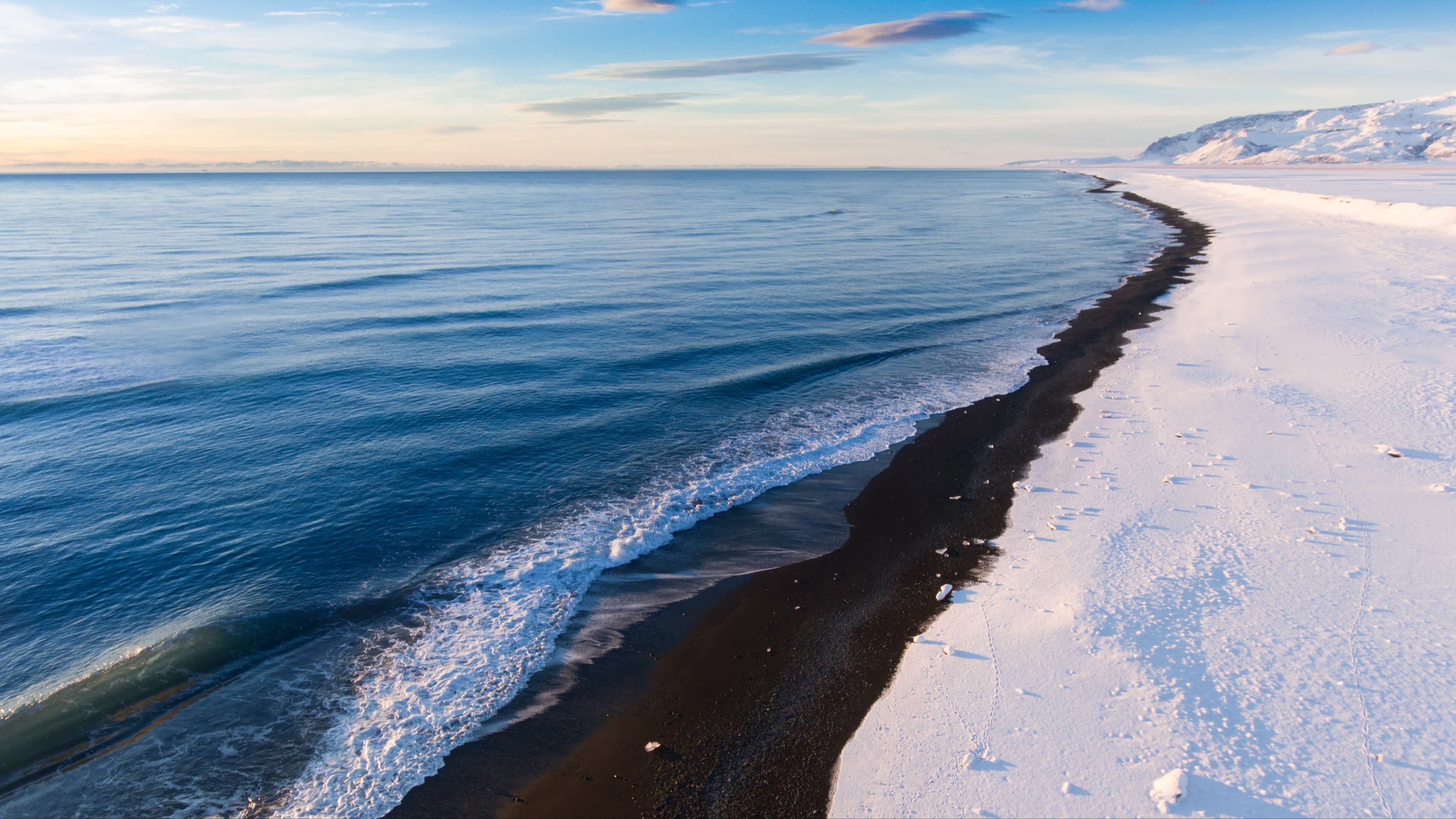 Winterlandschaft am schwarzen Sandvík-Strand in Island bei Sonnenschein und mit sanften Wellen im Meer. 
