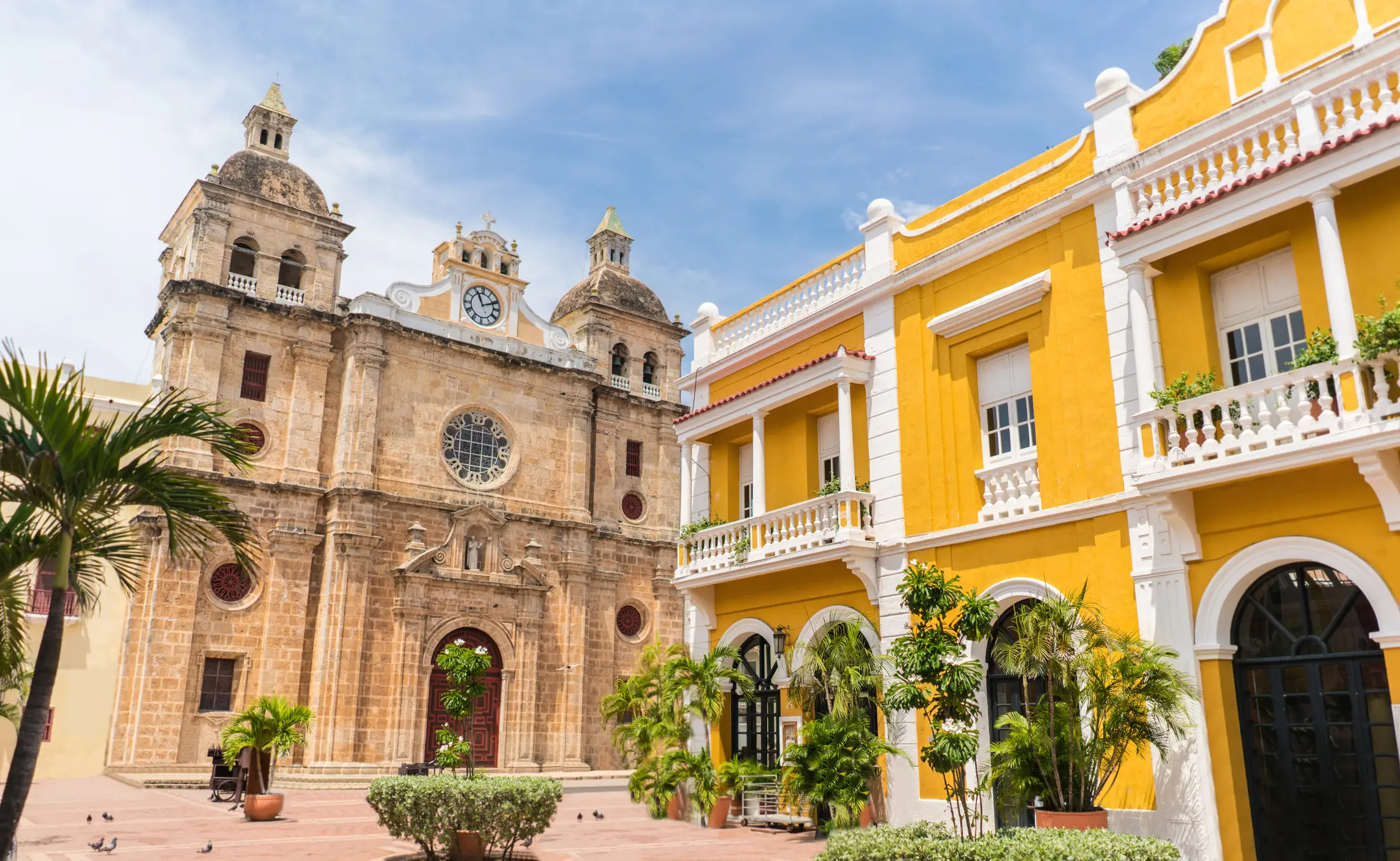 Historic stone church with bell towers next to vibrant yellow colonial building with white balconies, palm trees in foreground.