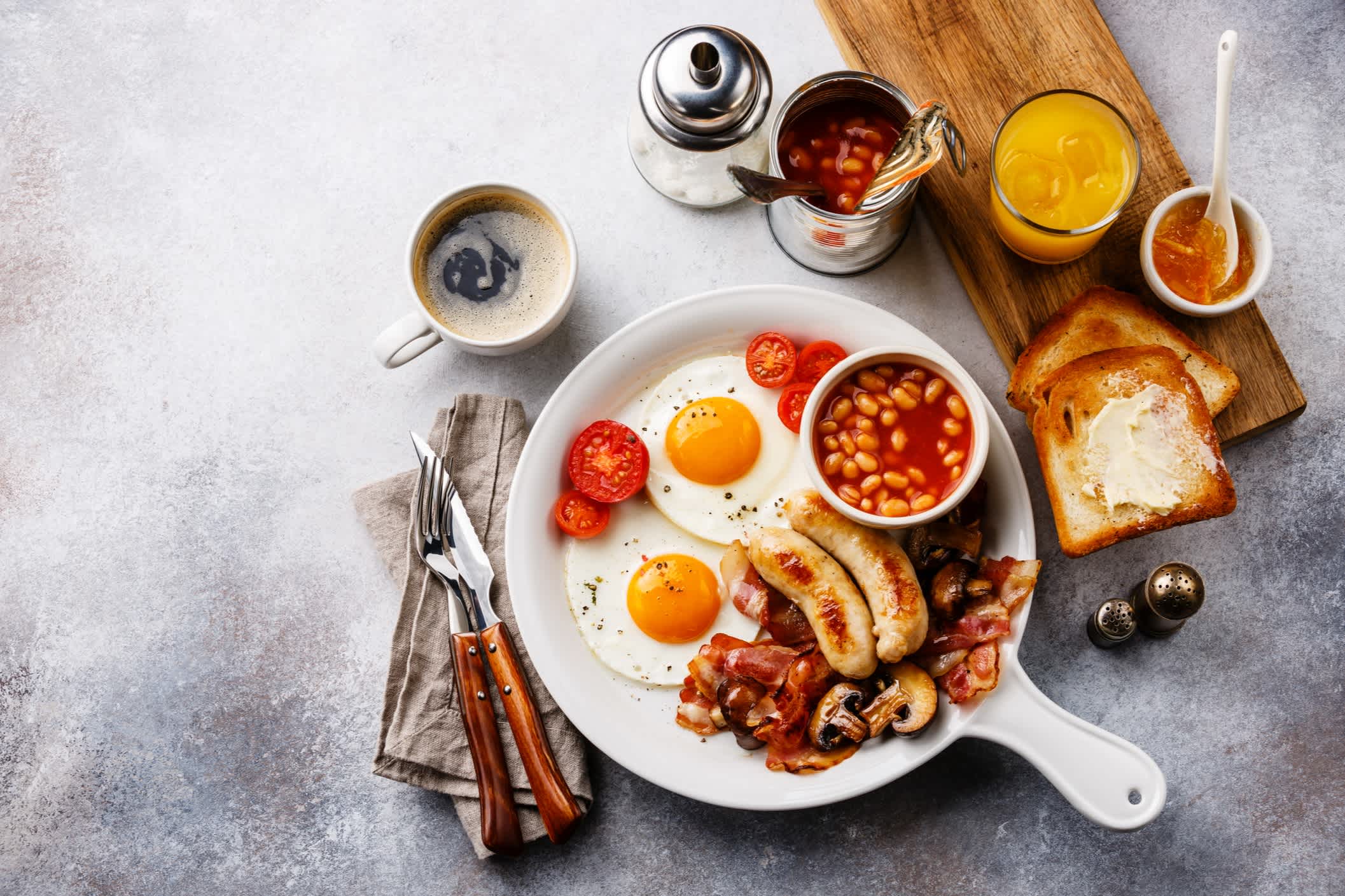 Englisches Frühstück in weißer Pfanne mit Spiegeleiern, Würstchen, Speck, Bohnen, Toast, Orangenfrisch und Kaffee auf weißem Hintergrund Kopierfläche