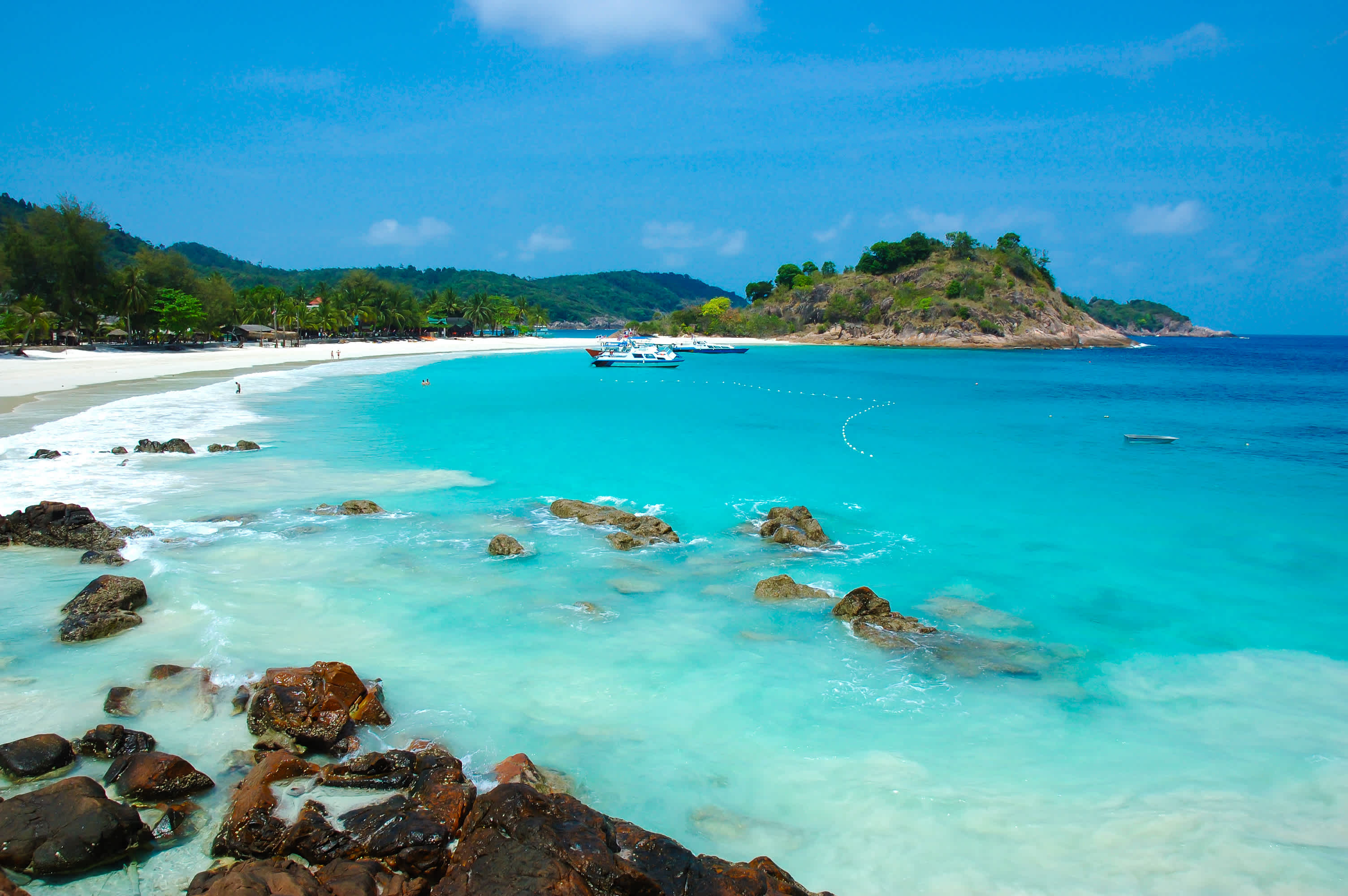 Rocher dans l'eau turquoise sur une plage de sable blanc avec bateaux en arrière-plan, Malaisie