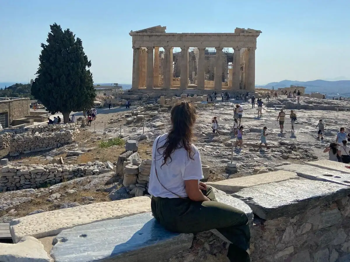 Viktoria assise contemplant le Parthénon à Athènes sous un ciel bleu, entourée de visiteurs et de ruines antiques.