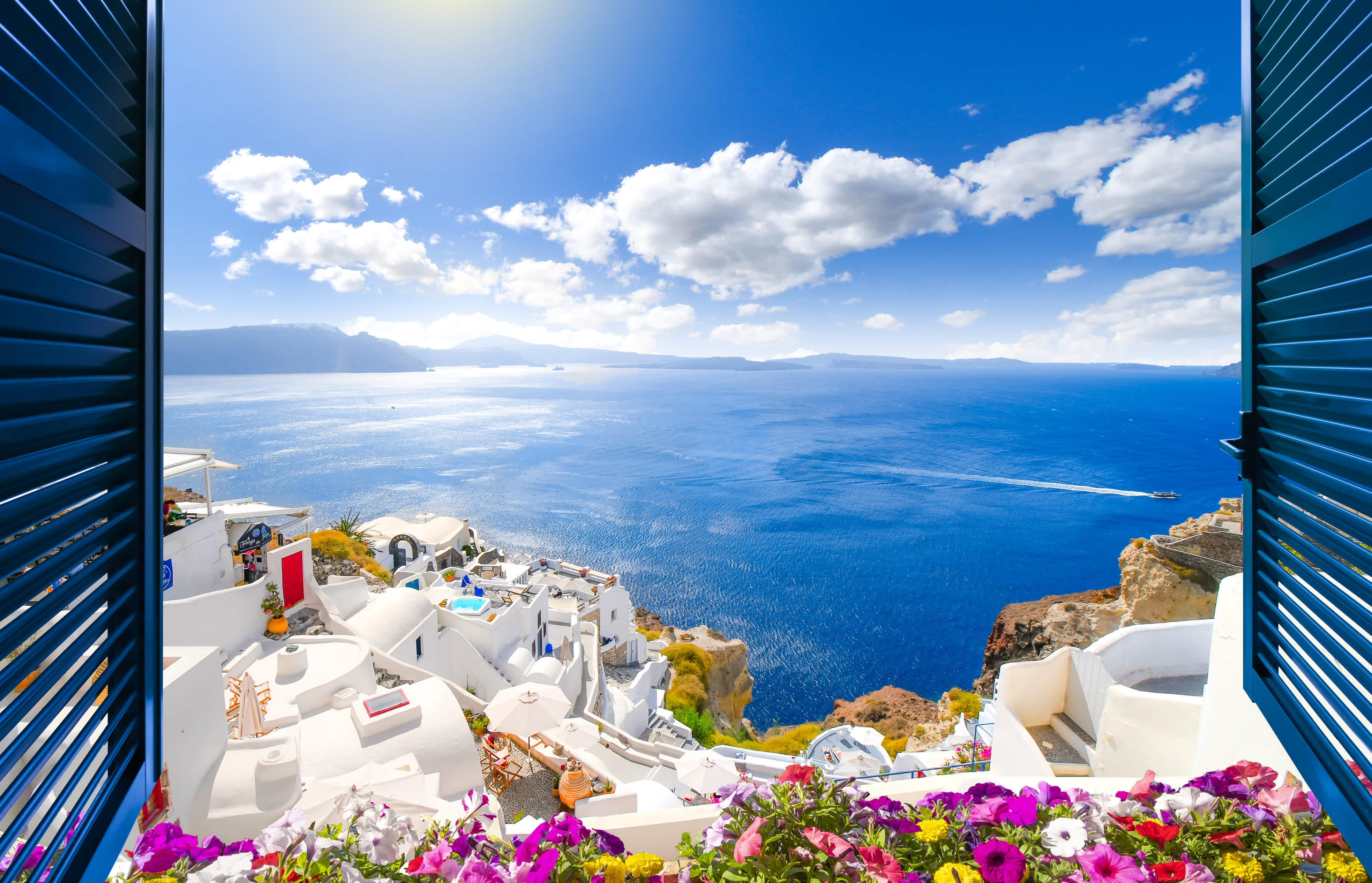 Vue à travers une fenêtre ouverte avec des volets bleus sur le village blanc d'Oia, qui se dresse au-dessus du bleu de la mer Égée sur l'île de Santorin, en Grèce.