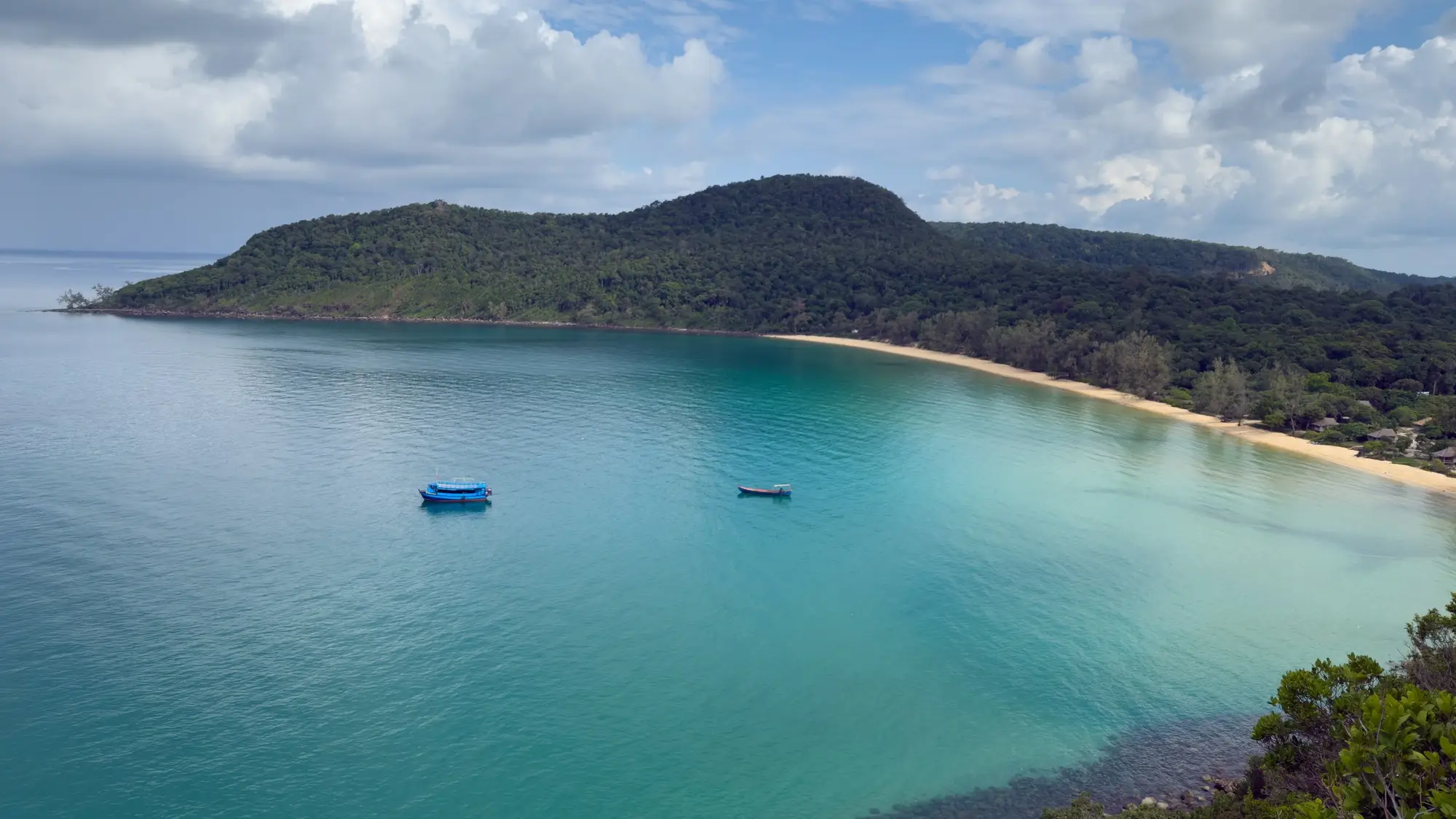 Tropische Bucht mit türkisblauem Wasser, zwei kleinen Booten und bewaldeter Insel unter teilweise bewölktem Himmel.