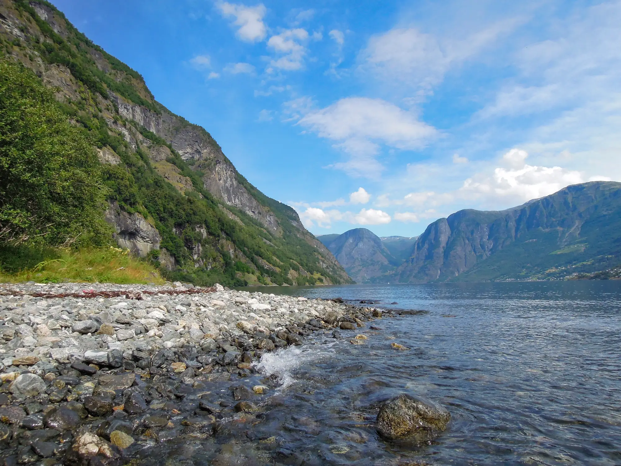 Eine felsige Küste mit klarem Wasser, grünen Hügeln und hohen Bergen unter einem blauen Himmel mit vereinzelten Wolken im Hintergrund.