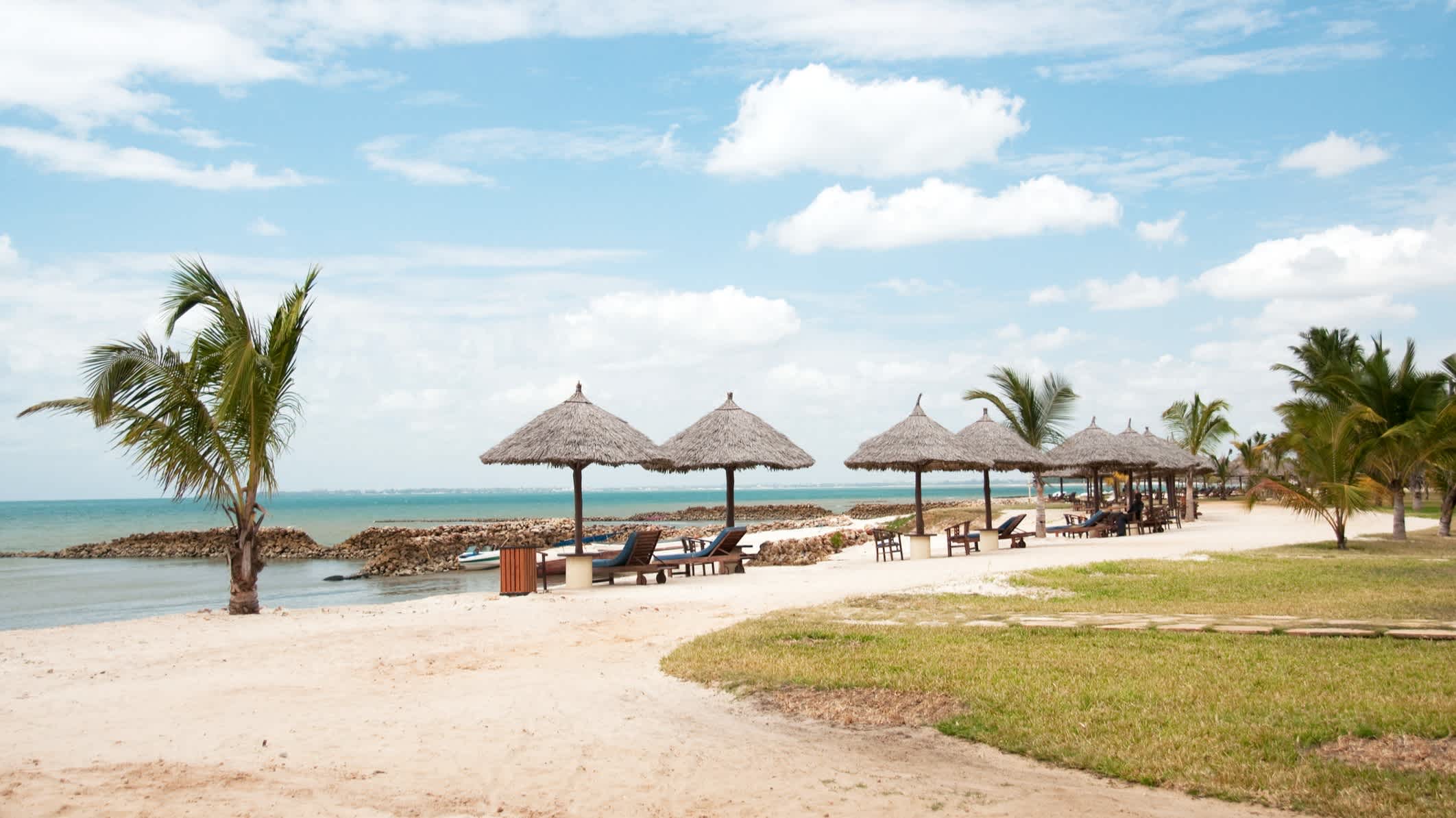 Sandstrand mit hellblauem Meer und Strohsonnenschirmen sowie Strandliegen und Palmen in der Nähe von Dar es Salaam, Tansania.